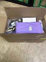 A top view of a large cardboard box containing a purple plastic-wrapped package, electrical cords, and other small items, mostly packed and some loose.