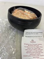 Black metal singing bowl with engraved patterns containing wrapped accessories inside, resting on bubble wrap.