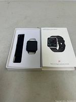 Photo of open box showing the black square smartwatch and extra black strap inside white package with product image showing on the box lid