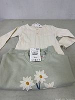 Two Zara kids tops displayed; cream long sleeve ribbed top and green floral sweatshirt, both size 3 with tags attached.