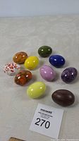 Photo of ten colorful ceramic eggs arranged on a light patterned fabric surface showing various solid and speckled colors.