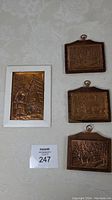 Photo showing all four copper embossed plaques on wood arranged on a flat surface, three smaller natural wood plaques on right and one larger framed white wood plaque on left