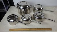 Overview of the five stainless steel cooking pots with lids, arranged on a table next to a ruler for size reference.