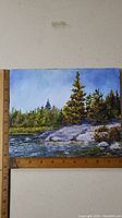 Oil painting on board of a river or lake shoreline with rocks and evergreen trees under a blue sky, shown with ruler measuring size.