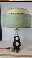 Full view of the lamp showing the green textured outer lampshade with stitching and the cream inner lampshade with stitching, lamp turned off.