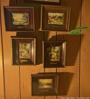 Five framed prints mounted on wood panel surface showing various cottage and village scenes. Each print has a brass plaque with title.