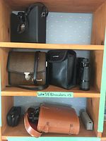Seven sets of binoculars on wooden shelves with cases and straps visible. Variety of sizes and materials.