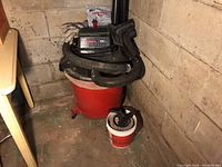 Red and black Craftsman 60L shop vacuum with attached hose and accessories on the floor next to a white and red bucket containing vacuum attachments, along with men's work gloves on top of the vacuum and convenience vacuum bags in original packaging behind.