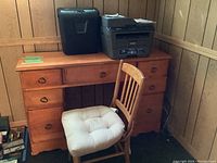 Desk and chair with cushion, Brother printer and Staples shredder on desk against wood-paneled wall.