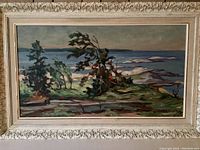 Full shot of the framed oil painting showing landscape with trees, water, and rocks under a cloudy sky. Painted with broad brushstrokes in greens and blues.