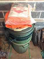 Two green plastic garbage cans stacked with two unopened polyethylene tarps on top, set against a brick wall in a garage.