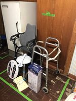 Photo showing grey commode chair, two aluminum walkers, handheld shower wand, footrest scale, and heating pad.
