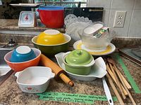 Four colorful Pyrex nesting bowls (blue, orange, yellow, green), ceramic baking dish with green lid, glass salad bowl, wooden spoons, and yellow and white concentric circle pedestal cake plate on kitchen counter, with paper doilies behind