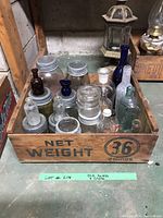 Photo showing a wooden crate containing a variety of old glass bottles and jars, some with metal lids, in assorted colors such as clear, brown, cobalt blue, and aqua green.