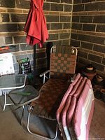 Photo showing red sun umbrella with stand behind a brown-green woven strap patio lounger. Four folded pink padded chair cushions placed on the lounger. Two small outdoor side tables, one square and one rectangular glass top.