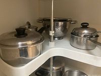 Corner cabinet shelf with several pots with lids and stainless steel strainers stacked.
