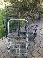 Three folding shopping buggies: two large dark blue and one smaller silver, outdoors on paved ground next to shrubbery