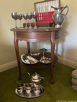 Photo showing silver plated goblets, water pitcher, and candy trays displayed on wooden table with shelf