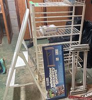 White metal wire storage rack with four wire shelves, step ladder next to it, boxed window security guards leaning against rack base, roof de-icing cable partially visible in rack basket.