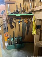 Photo of woodworking hand saws and a green metal toolbox with files and chisels beneath a shelf.