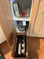 Baking pans stacked in a cardboard box inside a kitchen cabinet along with a glass casserole dish, two glass measuring cups with red lids, a metal box grater, and a hardcover cookbook.