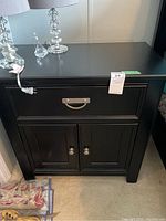 Front view of the black wood bedside table with one drawer and two doors below, showing silver handles and the matte finish.