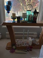 Front view showing assortment of candles and candle holders on wooden stand by window including tall blue glass holders, black metal lantern, turquoise glass lantern, and various candles.