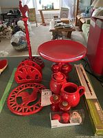 Full overview of the red cast iron kitchenware lot featuring paper towel holder, cake plate, cream and sugar set, salt and pepper shakers, napkin holder, trivet, and porcelain olive dish on table.