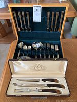Full view of stainless steel cutlery set arranged inside wooden case with lids open, containing spoons, forks, knives, and ladle.