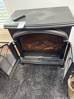 Front view of the black electric fireplace with both doors open, showing faux logs inside and the inner panel with control buttons.
