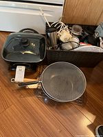 Photo showing electric fry pan with glass lid and various kitchen items including cooling racks, mugs, flashlight, and containers in a basket.