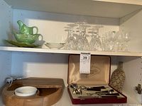 Top shelf with green cream and sugar set, crystal wine glasses, and liqueur glasses, wooden cheese board, bar accessories and decorative bottle.