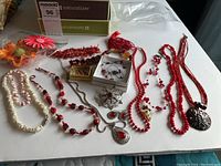 Photo shows a collection of fashion jewellery including several red bead necklaces, a brooch marked sterling with some missing gems, and various other necklaces with white and natural colored beads.