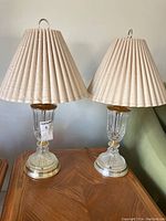 Pair of glass lamps with beige pleated fabric lampshades, ribbed glass bodies, and brass-tone metal bases on a wooden surface.