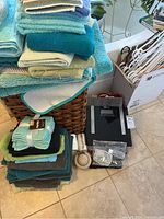 Wicker laundry basket with stacks of assorted towels in shades of blue, green, and brown, a digital scale, and various other household items including brushes and cords.