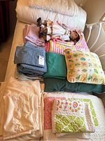 Photo showing multiple folded linens including sheets in cream, light blue, teal, gray, quilts with floral and patchwork designs, yellow pillow with animals, and a doll lying on top.