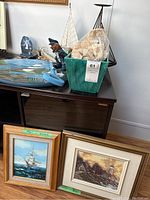 Framed paintings leaning on wall showing sailing ship at sea and lighthouse scene, along with nautical figurines and netted basket on cabinet top.