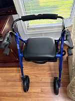 Front view of blue walker rollator with black padded seat and hand brakes on wooden floor near window.