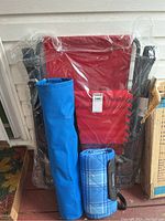 Red zero gravity relaxer folded and wrapped in plastic, accompanied by a blue carrying bag and blue plaid blanket rolled and strapped.