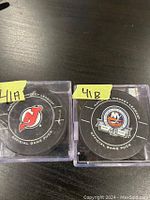 Front view of two official NHL game used hockey pucks, one New Jersey Devils and one New York Islanders, each in a clear plastic display case on a wooden surface.