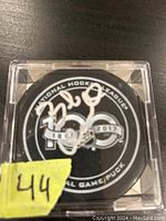 NHL 100 Greatest Game puck signed by Bobby Clarke, displayed inside a clear plastic case with a yellow lot number sticker.