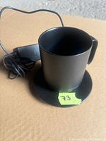 Top view of black Ember coffee mug placed on charging base with attached power cord, showing mug interior and base surface with sticker.