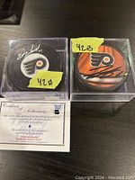 Two signed Philadelphia Flyers hockey pucks, one signed by Doug Favell and the other by Travis Sanheim, each in a protective clear display case with certificate of authenticity visible underneath one puck.
