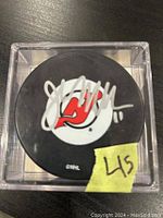 Front view of black New Jersey Devils hockey puck signed in white by John MacLean, housed inside clear plastic display case. Yellow sticker marked L45 visible.
