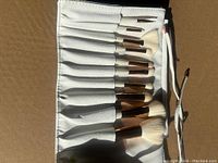 Set of 12 makeup brushes with white bristles and rose gold ferrules laid out in white leather pouch.