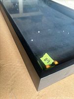 Close-up photo of corner of black shadow box with glass top and yellow sticker number 68 on inside corner, showing some dust spots and light surface smudges.