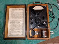 Full view of open wooden case showing flux meter device, attached cord, and inside lid with operating instructions.