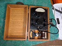 Wooden case opened showing the Marion TS 15A/AP Flux Meter device with dial meter labeled 'Gauss', control switches, and probe secured inside. Instruction sheet inside case lid. Leather strap attached but appears broken.