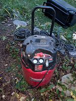 Husky air compressor showing control panel, pressure gauges, and handle, surrounded by garden area with hoses coiled nearby.