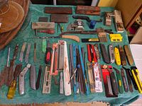 Full lot of blades and related cutting tools displayed on green surface, includes various utility knives, hacksaw blades, razor blade packs, sharpening stones, and hand tools.